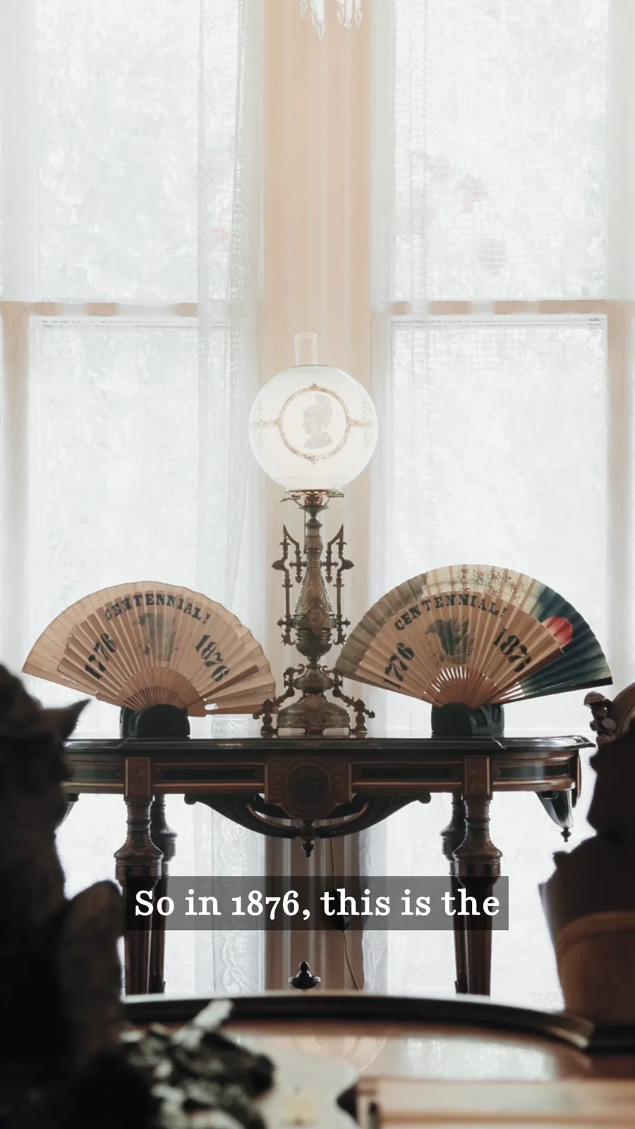 Two ornate folding fans displayed on a stand by a lace-curtained window